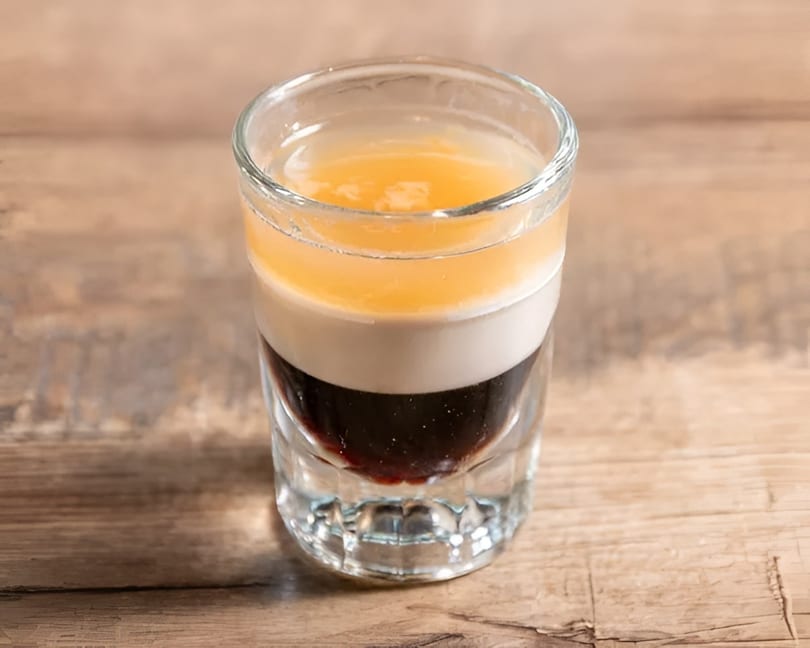 Layered shot glass with dark, white, and orange liquids on a wooden surface.