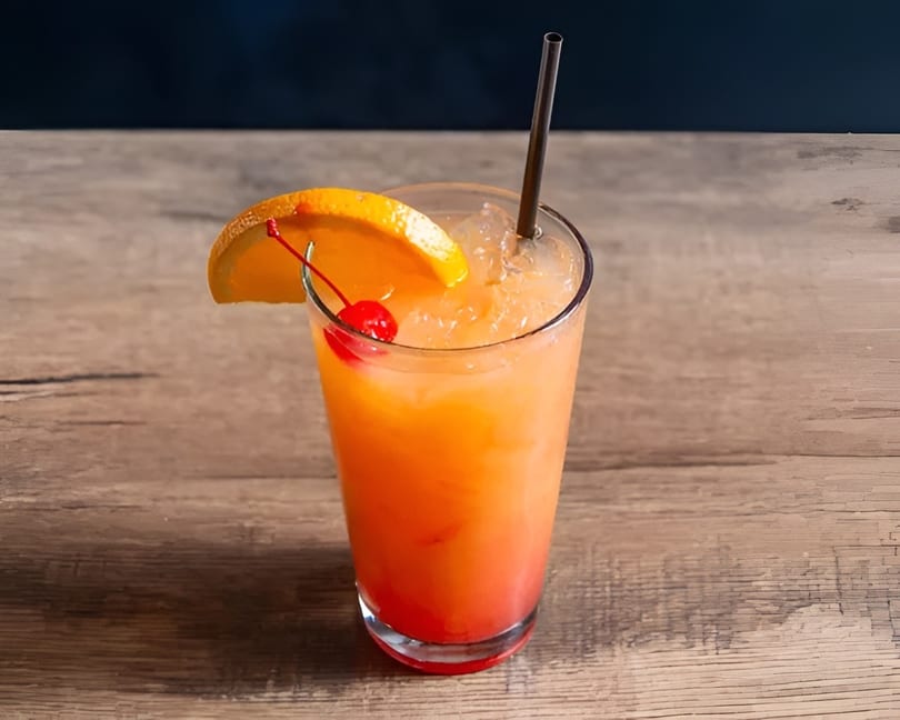A vibrant orange and red cocktail with an orange slice and cherry garnish, on a wooden table.