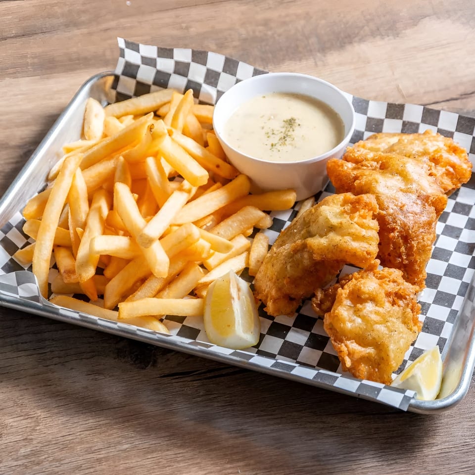 Golden fish and chips with tartar sauce and lemon wedges on a checkered paper-lined tray.