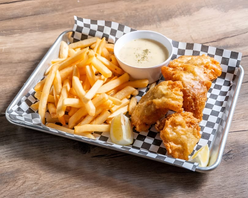 Golden fish and chips with tartar sauce and lemon wedges on a checkered paper-lined tray.