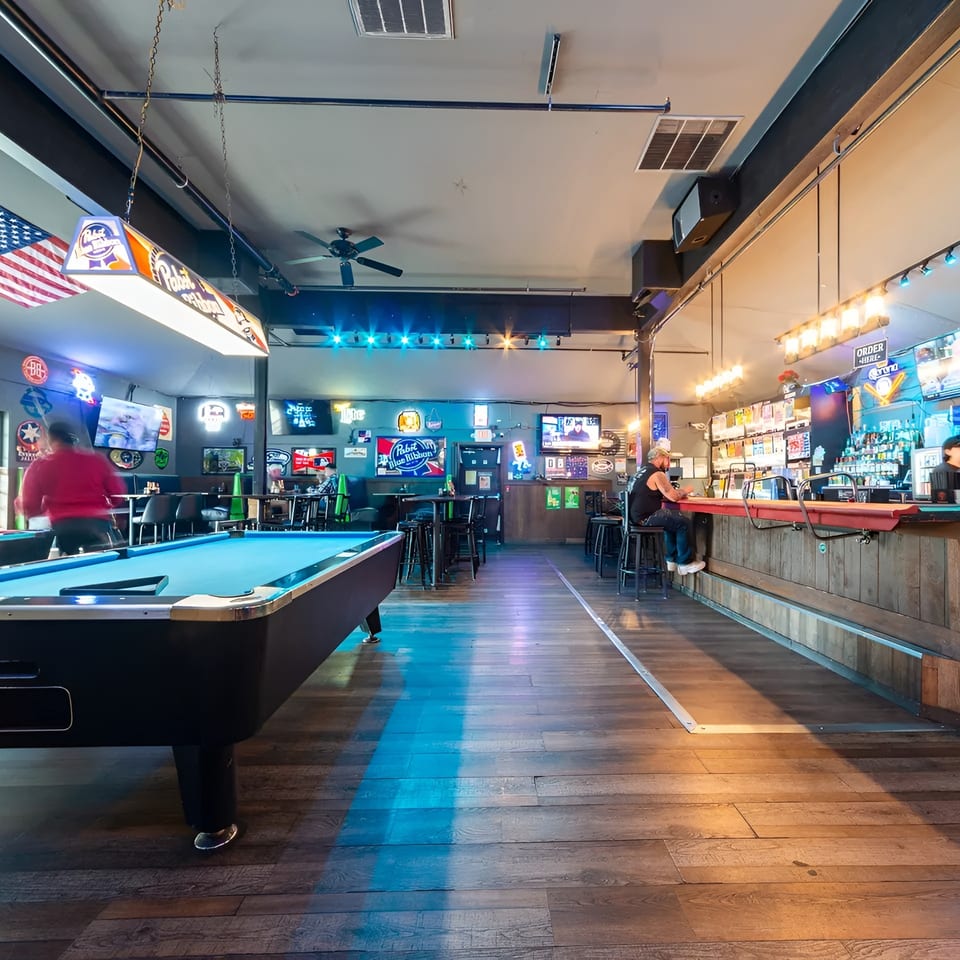 A lively bar interior with a pool table, patrons, and a well-stocked bar with neon signs.