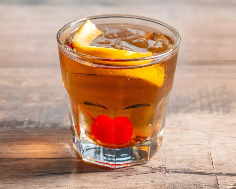 An Old Fashioned cocktail with a cherry and orange peel on a wooden table.
