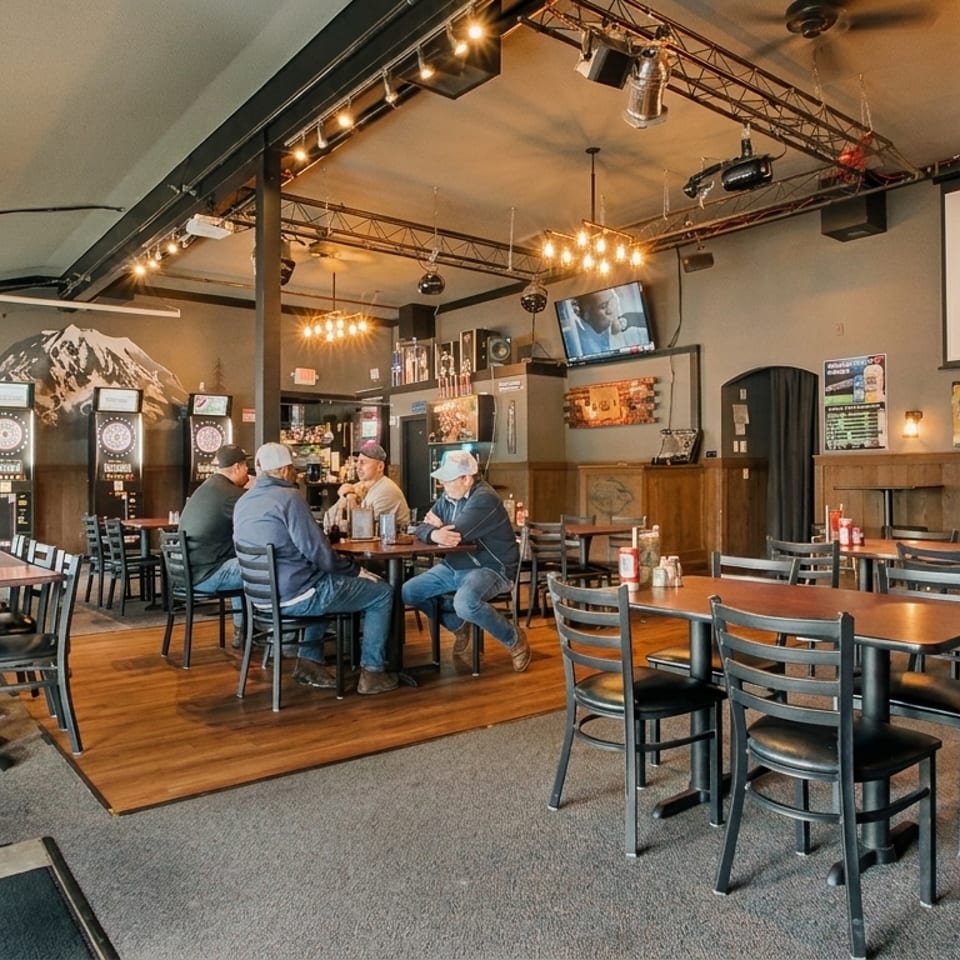 A casual restaurant interior with patrons at tables, dartboards, and a mountain mural on the wall.