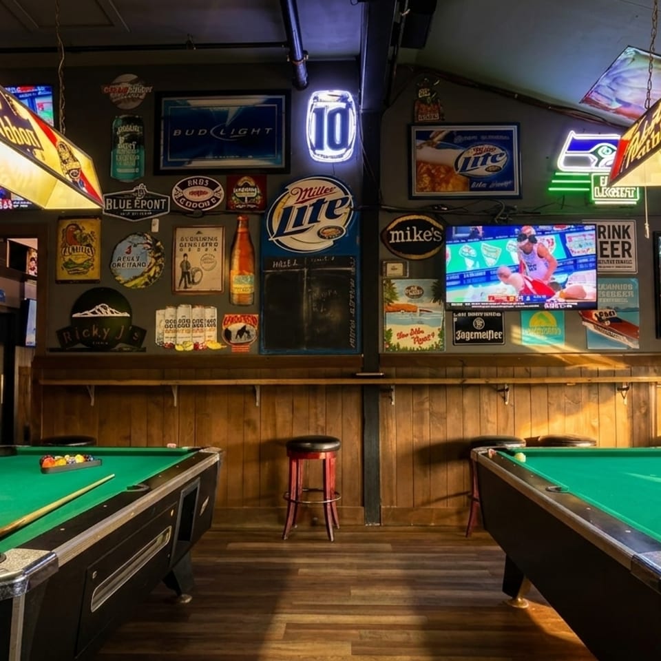 Two pool tables under beer-branded lights in a dimly lit bar with neon signs and TVs.