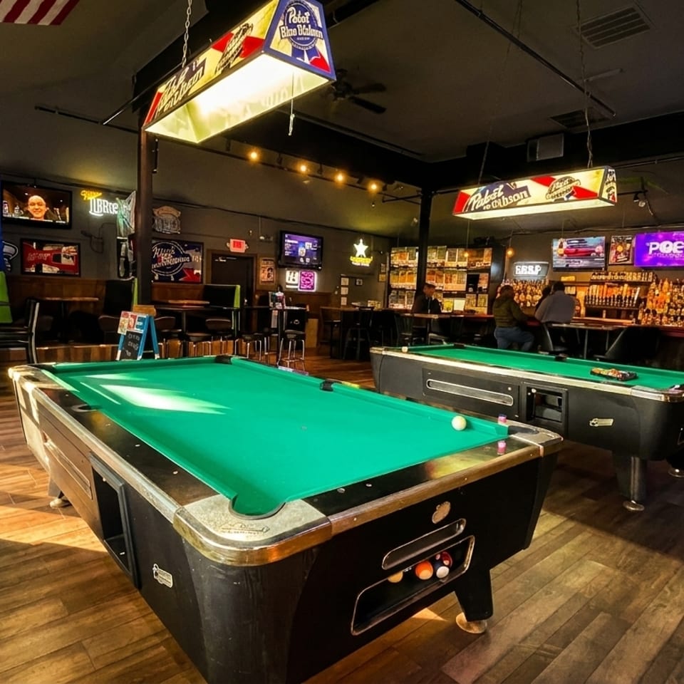 A lively bar interior with two pool tables, a long bar, and multiple TVs showing sports.